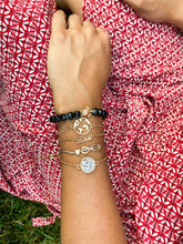 Load image into Gallery viewer,  Woman resting her wrist on her dress, wearing in writs 5 bracelets: one black with a turtle, one golden with a shape of the world, one golden spelling &quot;love&quot;, one with a heart and a symbol for infinite, and one golden with white stones
