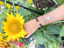 Load image into Gallery viewer, Hand holding a sunflower, wearing in writs 5 bracelets: one black with a turtle, one golden with a shape of the world, one golden spelling &quot;love&quot;, one with a heart and a symbol for infinite, and one golden with white stones
